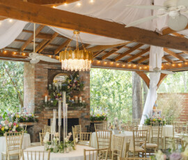 Chic outdoor wedding venue with white-draped tables, gold chairs, floral centerpieces, chandeliers, and brick fireplace.