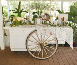 White cart with floral cloth, candles, photos, and greenery under pavilion; rustic decor for a Louisiana wedding.