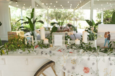 Wedding welcome table decorated with framed photos, candles, greenery, and floral linens.