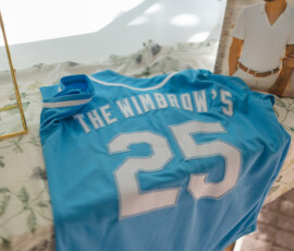 Light blue sports jersey with “THE WIMBROWS” and 25 on a floral tablecloth beside a wedding photo of a couple in white.