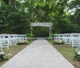 Outdoor wedding aisle with white chairs, colorful flowers, and floral archway under green trees in the Gulf South.