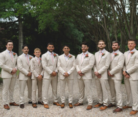 Ten men in beige suits with boutonnieres pose on a gravel path for a formal wedding photo, surrounded by Louisiana greenery.