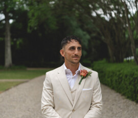 Man in beige suit with peach boutonniere stands on a gravel path amid greenery before a New Orleans wedding ceremony.