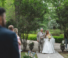 Bride in white gown walks down a garden aisle with an older man, surrounded by greenery and guests watching.