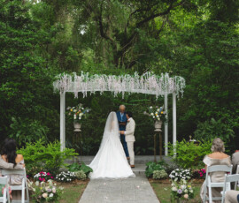 Bride and groom hold hands under a floral arch at an outdoor New Orleans wedding as guests watch from white chairs.