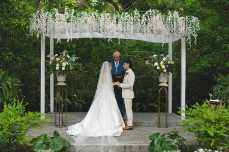 Outdoor garden wedding ceremony with couple exchanging vows beneath a white floral arbor.