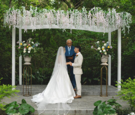 Bride and groom hold hands under a floral arch at their outdoor New Orleans wedding, surrounded by lush greenery.