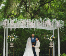 Couple weds under white floral arch with officiant in outdoor Louisiana ceremony, bride in long gown and veil, greenery around.