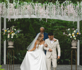 Bride and groom walk hand in hand under a floral arch during their New Orleans wedding, surrounded by lush greenery.