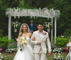 Bride and groom smile as they walk arm in arm down an outdoor aisle, surrounded by greenery at a classic Louisiana wedding.