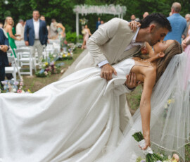 Groom in tan suit dips and kisses bride in white dress at outdoor Louisiana wedding as guests look on.