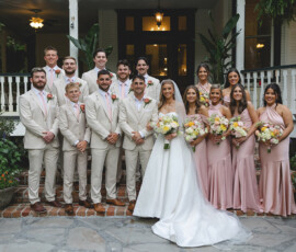 Bridal party in tan suits and blush dresses poses by a white house; bride in white gown holds bouquet at Louisiana wedding.