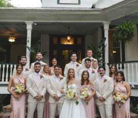Bride, groom, and wedding party pose smiling by a white house; bridesmaids in blush, groomsmen in light suits with pink ties.