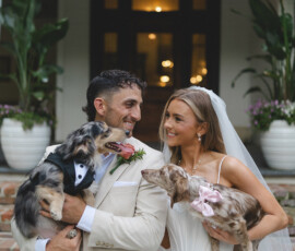 Bride and groom smile on brick steps, holding two small dogs in wedding attire, surrounded by plants and flowers.