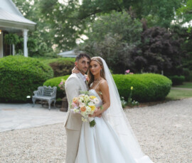 Bride in white gown and veil with groom in beige suit on a gravel path, greenery and white house behind—New Orleans wedding inspo.