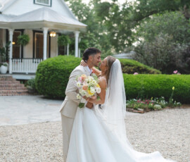 Bride and groom embrace and smile outside a white house, surrounded by greenery. Bride holds colorful bouquet in white dress and veil.
