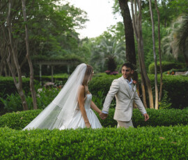 Bride in white dress and groom in light suit walk hand in hand through a lush, green Gulf South garden.