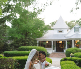 Groom in white suit carries bride in gown and veil through a lush garden at their New Orleans wedding, house behind them.