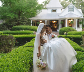 Groom in white suit carries bride in wedding dress and veil along a garden path by a large house, surrounded by greenery.