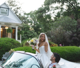Bride with flowers stands in vintage convertible, groom in light suit at wheel, both smiling by lush New Orleans home.