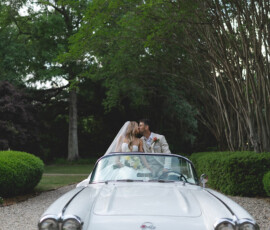 Bride and groom share a kiss in a white vintage Corvette on a gravel drive, surrounded by lush greenery.
