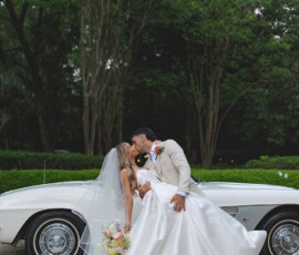 Bride and groom kiss by a white vintage car amid greenery; groom dips bride in long gown and veil, bouquet in hand.
