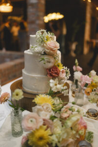 Three-tier white wedding cake decorated with fresh pastel flowers and greenery on a reception table.