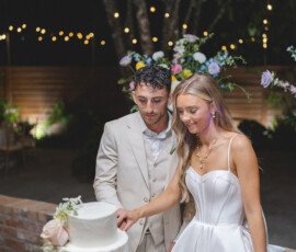Bride and groom cut a white tiered cake at an elegant Gulf South wedding, with flowers and glowing string lights in the background.