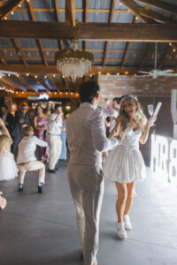 Wedding reception dance floor with couple dancing while guests celebrate beneath chandeliers and string lights.