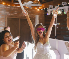 Two women dance at a lively New Orleans wedding, wearing white and pink dresses under string lights and large illuminated letters.