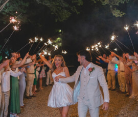 Newlyweds walk hand-in-hand under a tunnel of sparklers, bride in short white dress, celebrating a joyful Gulf South wedding.