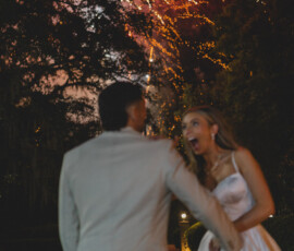 A couple in formalwear holds hands under colorful fireworks at night during their unforgettable Louisiana wedding reception.