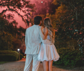 A couple in white watches fireworks illuminate the night sky above trees at their Louisiana wedding.
