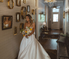 Bride in white gown with bouquet stands in elegant Louisiana hallway, sunlight streaming through door, creating a romantic scene.
