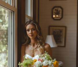 Bride in white dress holds colorful bouquet by a window in softly lit room, framed art on walls, New Orleans wedding inspo.