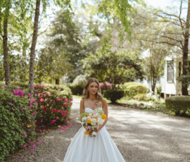Bride in white gown holding bouquet on a garden path with green trees and blooming bushes, perfect wedding inspiration.