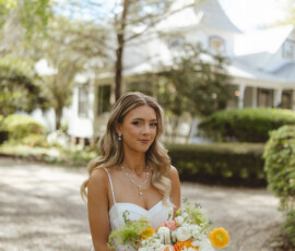 Bride in white dress holding colorful bouquet on gravel path, with greenery and white house, Louisiana wedding elegance.
