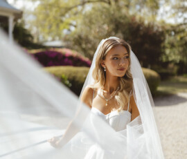 Bride in white dress and veil stands outdoors with bouquet, sunlight and greenery evoke Gulf South wedding romance.
