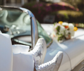 Feet in decorated white sneakers rest on a classic white convertible with flowers on the hood, perfect Gulf South wedding inspiration.