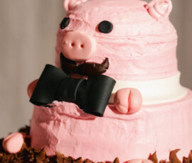 Whimsical pink groom’s cake shaped like a pig wearing a black bow tie with chocolate “mud” icing at the base.