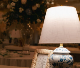 White and blue floral lamp beside framed invitation on white ledge, with blurred bouquet—Battle House Mobile wedding ambiance.