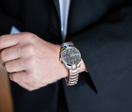 Person in black suit adjusting a silver, multi-dial wristwatch on left wrist; hands and part of suit visible.
