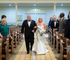 A bride in a white dress walks down the church aisle with her father, guests watching from wooden pews—Louisiana wedding inspiration.