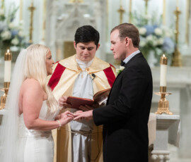 Bride and groom exchange rings in a Louisiana church wedding, with priest, candles, and floral altar arrangements.