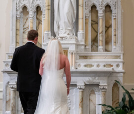Bride in white dress and groom in black suit stand before Virgin Mary statue at Louisiana church wedding.