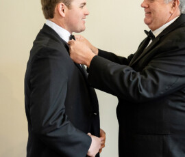 An older man helps a younger man with his bow tie; both smile in black tuxedos before a Louisiana wedding.