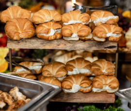 Croissant sandwiches on a two-tier wooden stand with fruits, buffet dishes, and a lit candle for a warm Gulf South wedding spread.