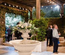 White fountain with flowers under string lights at an outdoor New Orleans-style wedding, guests chatting near a wooden pergola.