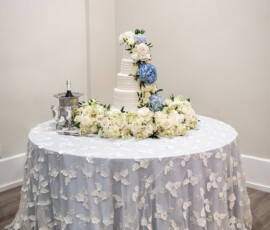 Round table with white lace cloth, tiered wedding cake with blue and white flowers, five bouquets, and a silver ice bucket.