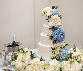 White tiered wedding cake with white and blue flowers, flower petals, silver goblets, and champagne—ideal for a New Orleans wedding.